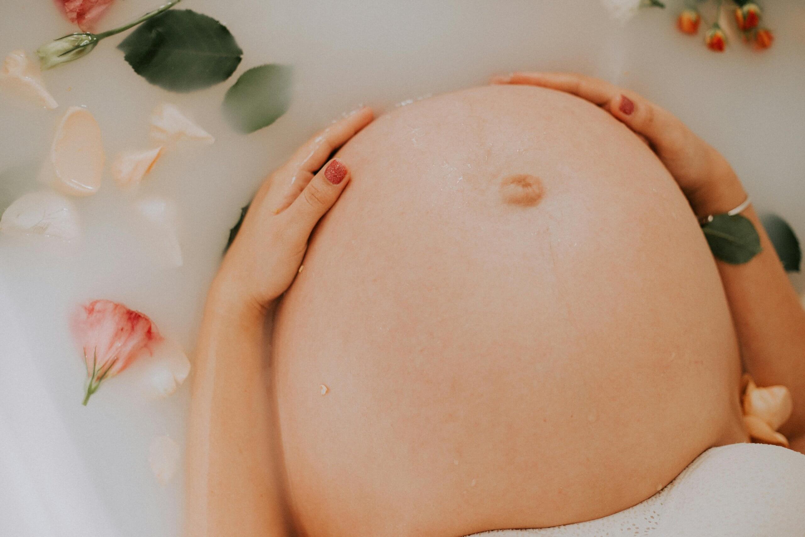 Femme enceinte se relaxant dans un bain de lait avec des pétales de fleurs, tenant son ventre arrondi avec douceur – préparation à l’accouchement en sérénité.