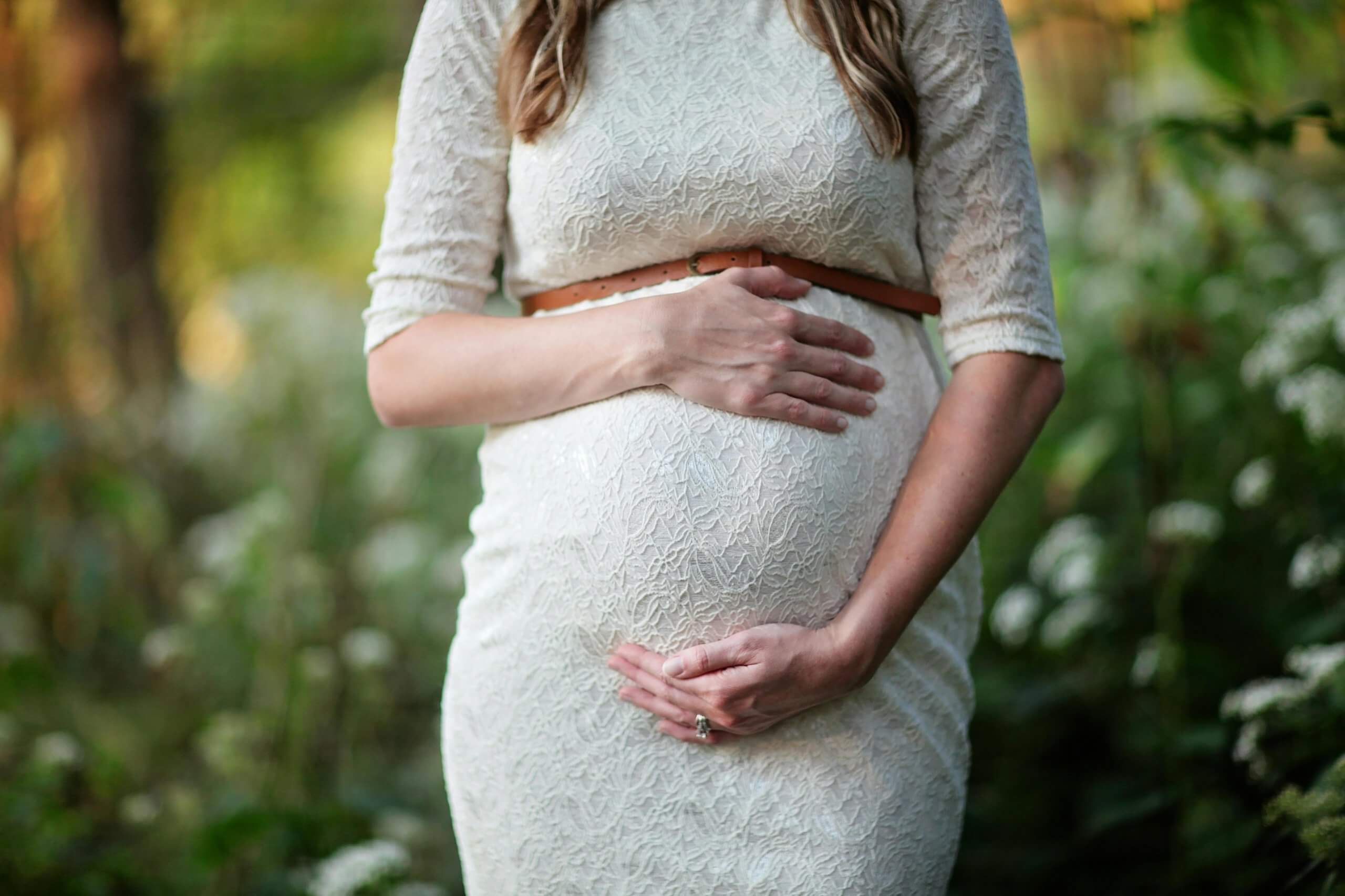 Femme enceinte en robe blanche tenant tendrement son ventre, illustrant la douceur de la grossesse et l’importance de prendre soin de son dos pendant cette période.