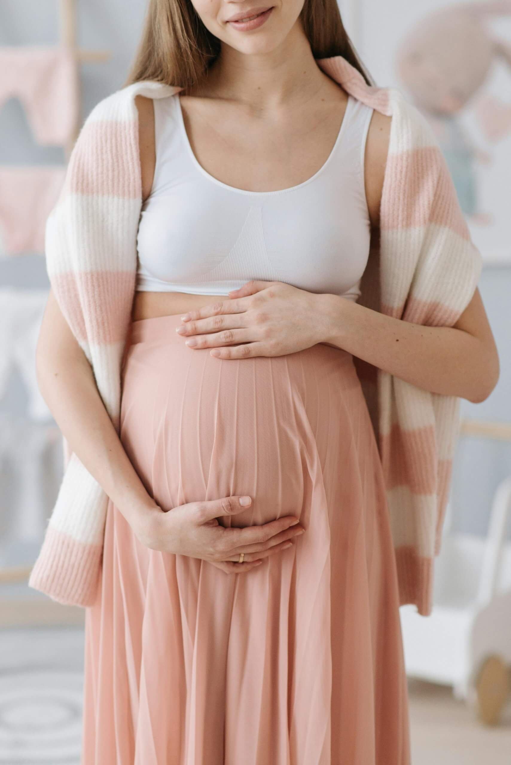 Femme enceinte habillée en rose et blanc, tenant son ventre. Cette image illustre l'importance de la gestion des douleurs de dos pendant la grossesse à travers des solutions naturelles comme l'ostéopathie, l'activité physique adaptée, et une bonne hydratation